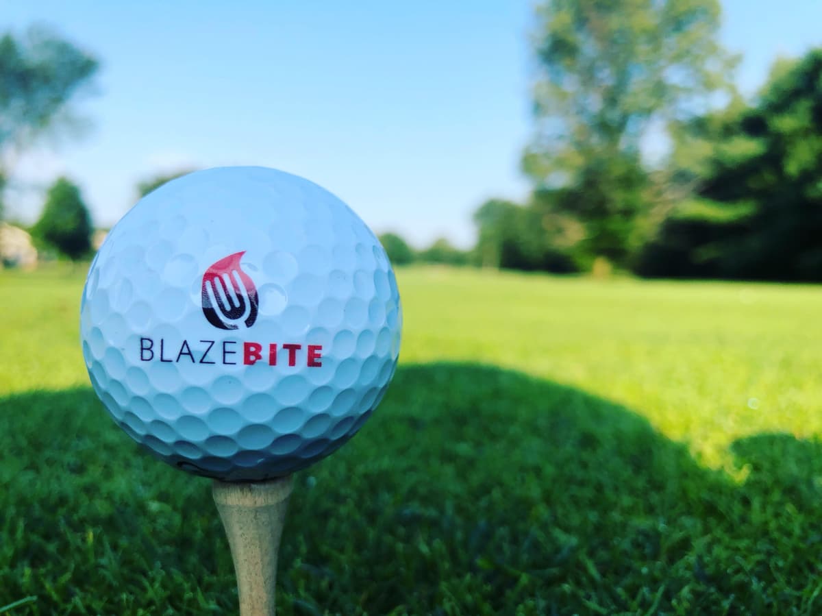 A golf ball printed with the BlazeBite logo on a tee at the edge of a fairway