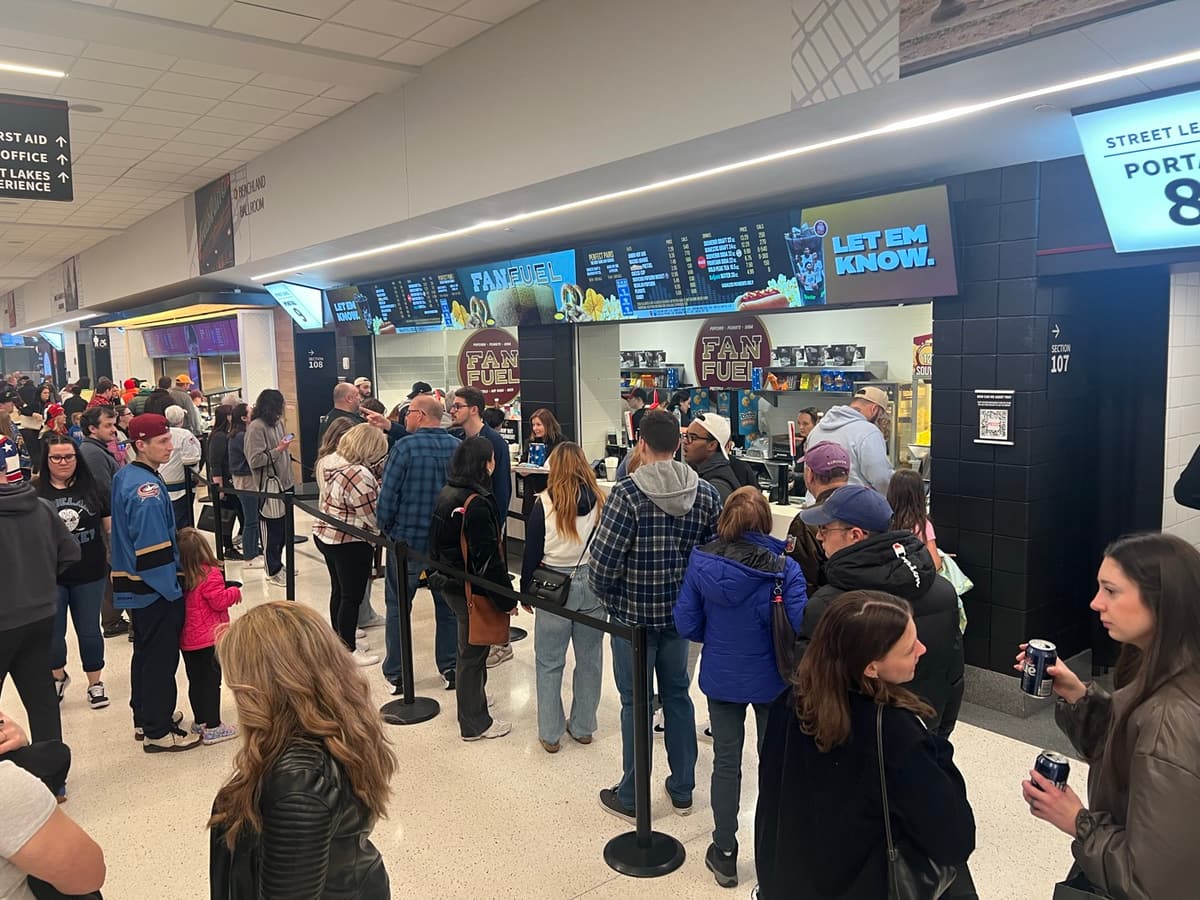 A crowd lined up at an arena concession stand between periods, with lit menu boards above the counter