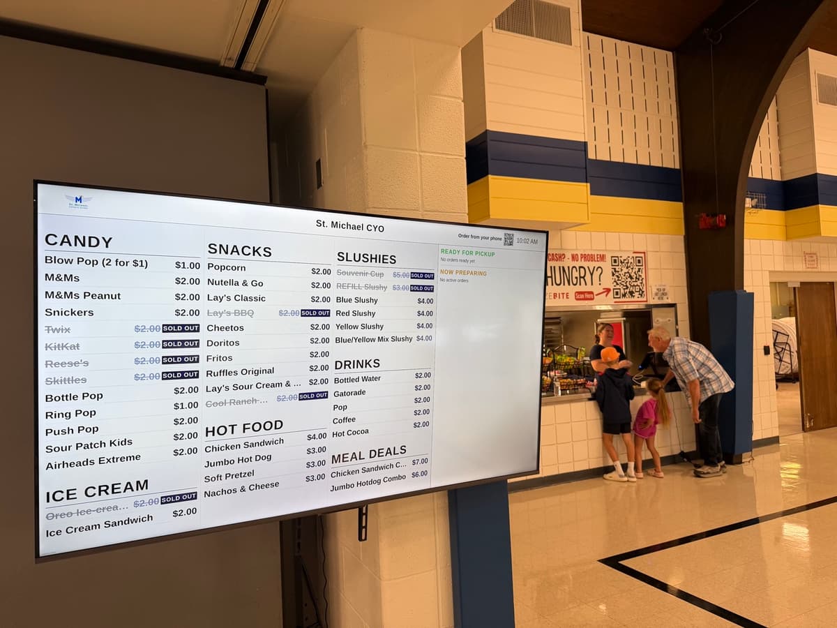 A BlazeBite menu board on a TV in a school lobby, with people at the concession counter