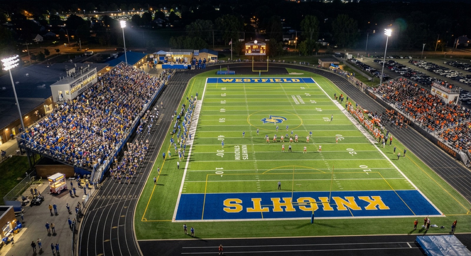 Aerial view of a packed high school football stadium at night, with a home grandstand, a visitor grandstand, and concession stands along the track