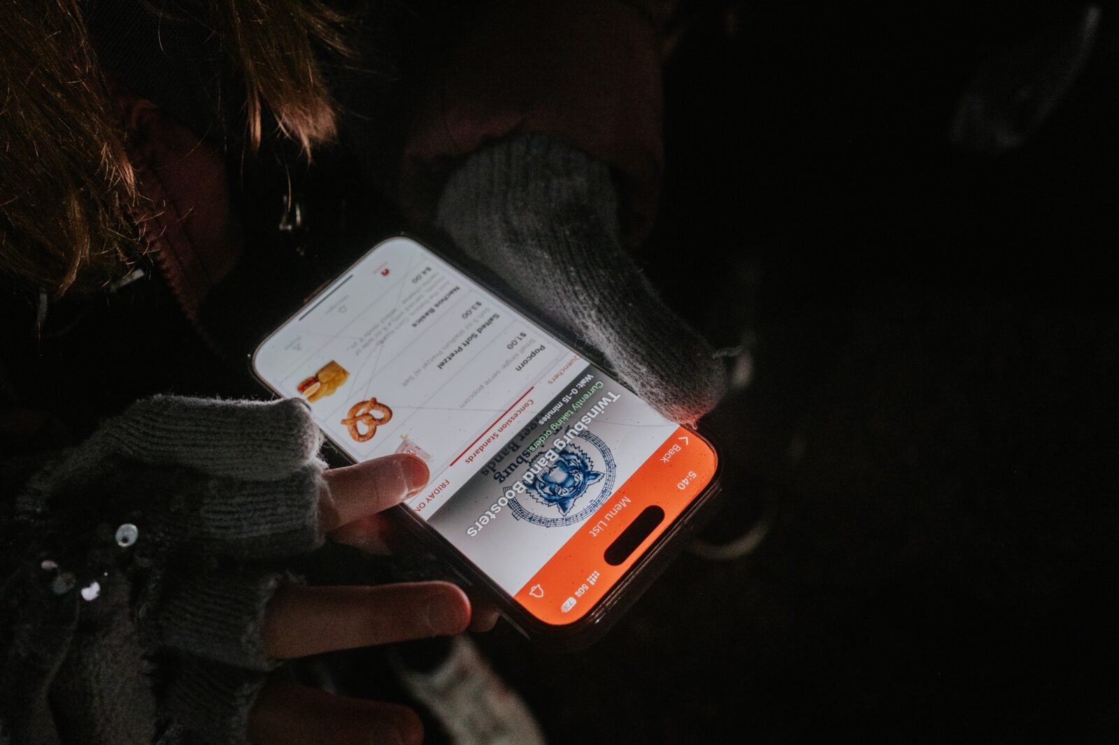 A fan in gloves and a winter coat holding a phone in the stands, browsing the Twinsburg Band Boosters concession menu on BlazeBite