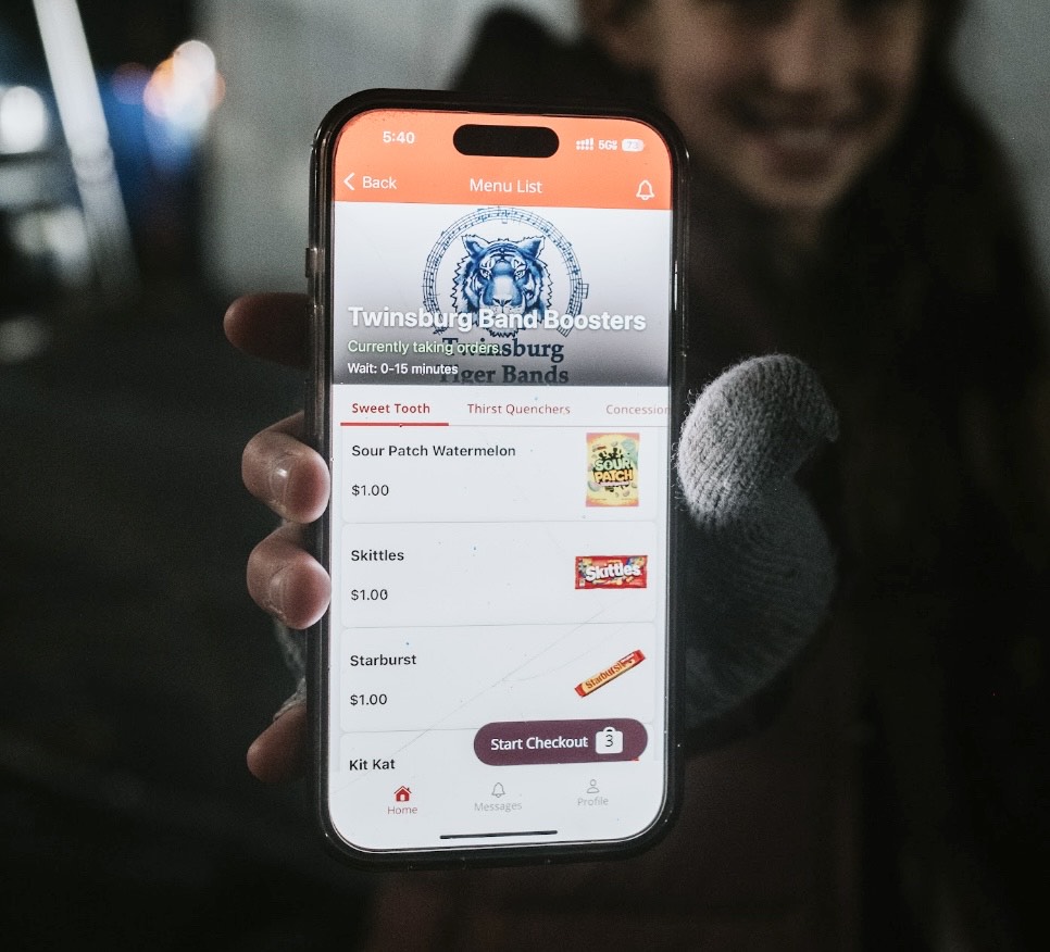 A hand holding a phone showing the Twinsburg Band Boosters menu in the BlazeBite app, with candy items and a Start Checkout button